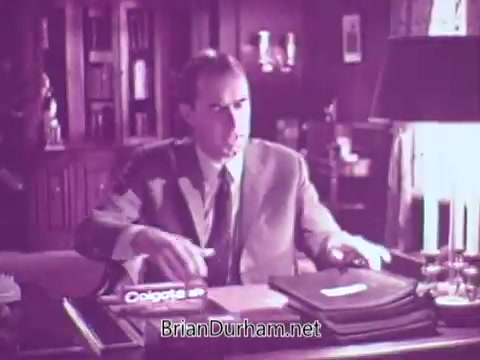 The still from the 1967 Colgate commercial features a man in a suit seated at a desk. The setting appears formal, with bookshelves and a lamp in the background. He is attentively engaging with something on the desk, which prominently displays the Colgate product, hinting at a presentation or discussion about the brand. The overall tone of the image is professional and focused.