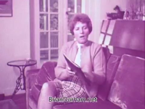 The 16mm still from the 1967 Colgate commercial features a woman sitting on a couch, dressed in a light cardigan and a patterned skirt. She is engaged in conversation, holding a magazine or pamphlet. The setting appears cozy, with a soft focus on the background, suggesting a home environment. The overall color tone is a muted pink, characteristic of the era.