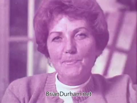 The still features a woman with short, styled hair and a light sweater, smiling while looking directly at the camera. The scene has a soft, pinkish hue, creating a nostalgic atmosphere typical of advertisements from the late 1960s. The background suggests a home setting with soft focus.