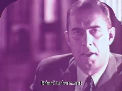 The still from the 1967 Colgate commercial features a man in a suit speaking directly to the camera. The image has a vintage aesthetic, with a purple tint, suggesting an advertising style typical of that era. The background appears blurred, focusing attention on the speaker, who likely promotes the benefits of Colgate products.