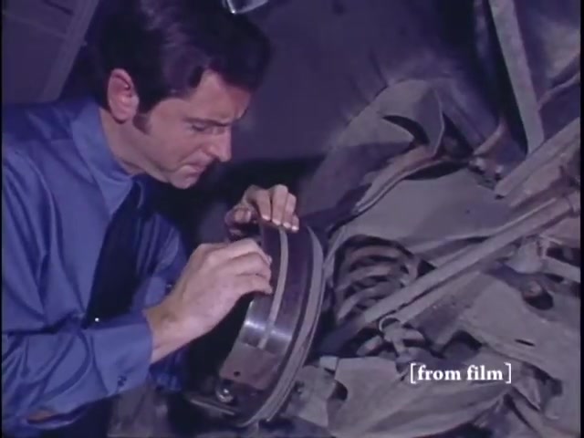 The still from the 'Nichols Automotive' commercial features a man in a blue shirt, closely inspecting a vehicle's brake component while working underneath a car. The environment suggests a garage or automotive shop setting. The visual quality reflects the characteristics of 16mm Ektachrome film, indicative of the era. The commercial concludes with the logo for Nichols Discount Center, emphasizing automotive services and customer care.