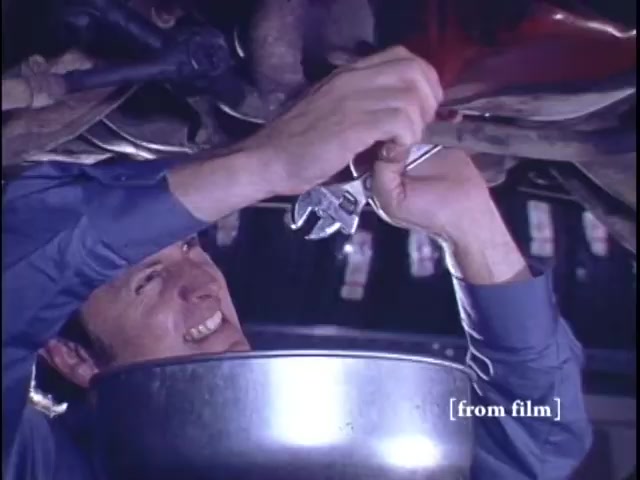 The still from the 1971 Nichols Automotive commercial depicts a mechanic working underneath a vehicle. He is smiling and using a wrench to perform a repair, with a metal basin positioned to catch any fluids. The scene conveys a sense of satisfaction and competence in automotive maintenance. The commercial's branding concludes with the Nichols Discount Center logo.