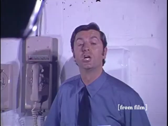 The still from the 'Nichols Automotive commercial - 1971' features a man in a blue shirt and tie speaking directly to the camera. He appears to be in an indoor setting, possibly an office or showroom, with a vintage telephone visible to his side. The image captures a moment from a commercial promoting Nichols Discount Center. The visual is reconstructed from Ektachrome film elements, and the lab paperwork confirms the 1971 date.
