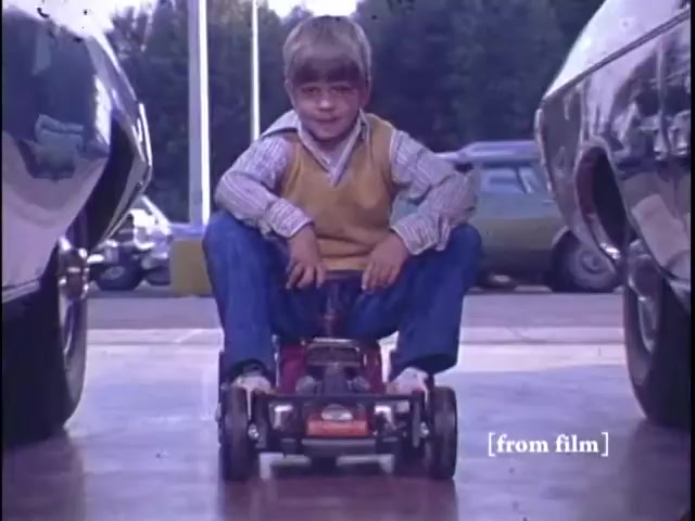 The still from the 1971 Nichols Automotive commercial features a young boy sitting on a toy car, with a playful expression. He is positioned between two larger vehicles in a car dealership, emphasizing the connection between childhood and automotive themes. The boy is dressed in a striped shirt and a vest, evoking a nostalgic, familial atmosphere. The scene captures a moment of joy and innocence, concluding with the logo for Nichols Discount Center.