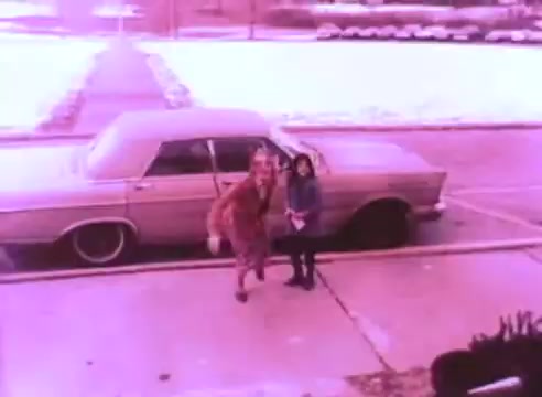 The still from 'PSA - Ex Offender 2' features a scene with a car parked on a snowy street. A woman in a coat and a child are seen outside, possibly interacting. The overall color tone is pinkish, giving the image a vintage look indicative of the film's era. The scene appears to highlight a moment of connection or community, relevant to the PSA's themes.