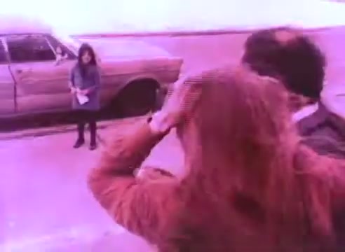The still from 'PSA - Ex Offender 2' depicts a street scene featuring a parked car and three individuals. A woman in the foreground, seemingly distressed, runs her hands through her hair, while another person stands beside her. In the background, a girl is separated from them, holding something in her hand, suggesting a moment of tension or communication. The overall color tone is muted, contributing to a serious atmosphere.