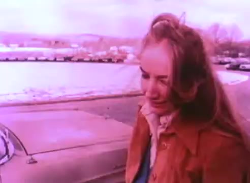 The still from 'PSA - Ex Offender 2' features a woman with long hair, dressed in a brown coat, standing outdoors near a parked car. The background shows a snowy landscape with vehicles and trees. The overall color tone appears to have a pinkish hue, suggesting a vintage or stylized aesthetic. The expression on her face seems contemplative or somber.