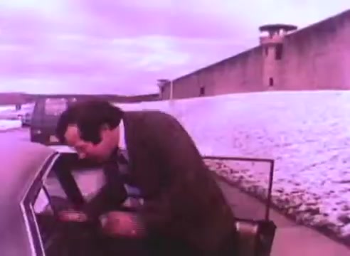 The still from 'PSA - Ex Offender 2' depicts a man in a suit leaning into a car near a snowy landscape. In the background, a large prison wall stands prominently, suggesting a context related to the challenges faced by ex-offenders. The overall tone appears somber, reflecting the themes of reintegration and societal perceptions.