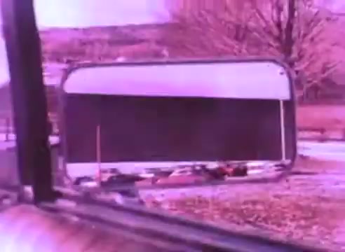 The still from 'PSA - Ex Offender 2' depicts a view through a vehicle's side mirror, showing a blurred reflection along with some indistinct objects and a background of trees. The dominant color is a soft purple hue, giving the scene an abstract quality. The image suggests themes of perspective and reflection, aligning with the public service announcement's focus on the experiences of ex-offenders.