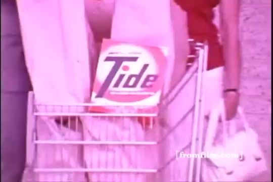 The still from the 'Tide TV Commercial - 1970s' features a shopping cart prominently displaying a box of Tide laundry detergent. The cart is positioned next to individuals dressed in casual clothing, suggesting a typical grocery shopping scene of the era. The overall color tone is slightly faded, typical of a 16mm print, giving it a vintage feel. The Tide logo is bold and recognizable, emphasizing the product's branding.