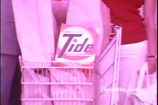 The still features a shopping cart filled with paper bags, prominently displaying a box of Tide laundry detergent. The scene captures a nostalgic 1970s aesthetic, with soft colors and a casual shopping environment. A person dressed in white pants and another in a red shirt are partially visible, enhancing the vintage feel of the commercial.