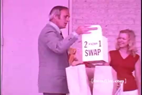 The still features a man in a gray suit and a woman in a red dress, both standing in front of a building. The man is holding a large carton displaying '2 for 1 SWAP,' likely promoting a Tide detergent offer. The scene has a vintage aesthetic typical of 1970s commercials, with a light pink hue dominating the image. The overall vibe is upbeat and promotional, typical of advertising from that era.