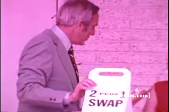 The still shows a man in a light grey suit holding a sign that reads '2 FOR 1 SWAP.' He appears to be engaged in a conversation or promotion. The background features a textured wall, creating a somewhat retro ambiance typical of 1970s commercials. The color tone is slightly faded, characteristic of a 16mm print.