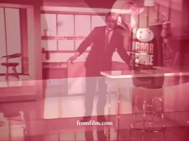 The still features a man in a suit gesturing enthusiastically in a bright, retro kitchen setting. The colors have a pinkish tint, likely a result of the 16mm print. Notable cleaning products, Glamolene, Liquid Glory, and Spray-n-Vac, are likely highlighted, presenting a classic advertising style. The overall aesthetic is nostalgic, reflecting the mid-20th century approach to promoting household cleaning items.