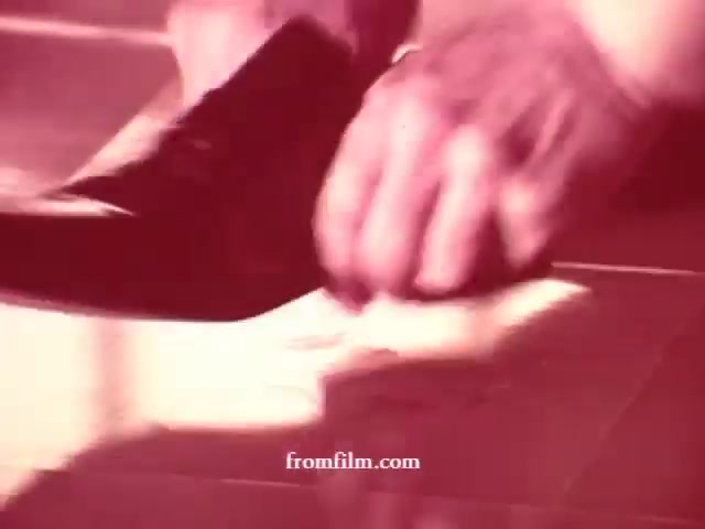 The still from the 16mm film features a close-up shot of a hand cleaning a surface, possibly a floor or countertop, with a cleaning product. A black shoe is visible in the frame, indicative of someone actively engaging in cleaning. The overall aesthetic appears vintage, reflecting the era of the commercials for Glamolene, Liquid Glory, and Spray-n-Vac. The colors seem slightly muted, characteristic of 16mm film quality, enhancing the nostalgic vibe of the cleaning product advertisements.
