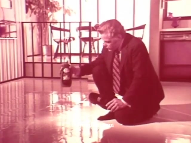 The still features a man in a suit, squatting on the shiny floor, applying a cleaning product from a bottle labeled 'Liquid Glory.' The setting appears to be a stylish, modern kitchen with a contemporary design. Natural light filters through, creating vibrant reflections on the floor. The image’s color palette evokes a vintage feel, typical of 16mm prints, enhancing the nostalgic vibe of the cleaning product promotion. The overall scene emphasizes the effectiveness and appeal of the cleaning product in a home environment.
