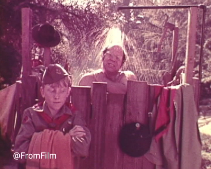 The still depicts a scene from a 1970s Band-Aid TV commercial, showcasing a humorous outdoor shower setting. A young boy, wearing a scout uniform and holding towels, stands next to a wooden enclosure where a man is taking a shower. The man appears jovial, with water cascading over him, while the boy looks on, likely amused or surprised. The colors are soft with a vintage feel, characteristic of 16mm film. The playful scenario highlights the everyday use and necessity of Band-Aids in a lighthearted context.