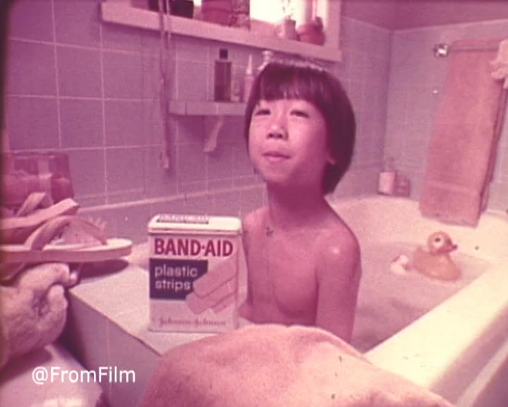 The still features a young child in a bathtub, smiling and holding a box of Band-Aid plastic strips. The bathroom has a vintage aesthetic, with a blue tiled background and soft lighting that creates a warm atmosphere. Accessories like a rubber duck and towels add to the playful setting, while the focus on the Band-Aid product suggests it's pivotal to the advertisement's message. The overall vibe is nostalgic, characteristic of 1970s commercials.