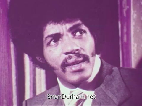 The still from the 16mm newsfilm 'Fight Against Record Piracy' from Stax Records captures a moment featuring a man with a distinct Afro hairstyle and a well-tailored suit, expressing concern or contemplation. The film discusses the issue of bootleg recordings rather than the anticipated cassette tape piracy, highlighting the impact of unauthorized copies on artists and the music industry in 1971. Overall, it reflects the serious efforts by Stax Records to combat the unauthorized reproduction of their music, emphasizing the importance of copyright protection in the creative sector.