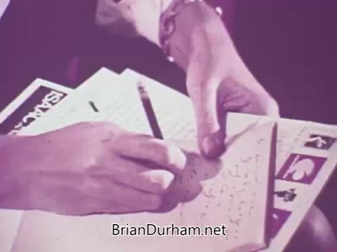 The still from the 16mm newsfilm 'Fight Against Record Piracy' from Stax Records captures a close-up of a hand writing notes in a notebook. The scene likely depicts a discussion or investigation into the issue of record bootlegging rather than amateur recordings. Papers with the Stax Records branding are visible, emphasizing the record label's concern over unauthorized reproductions of their music. This captures the essence of the era’s struggle against music piracy and the impact on artists and the industry.