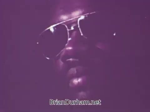 The still from the 1971 newsfilm 'Fight Against Record Piracy' highlights the serious issue of music bootlegging. It features a close-up shot of a man wearing sunglasses, set against a purple background, which adds a dramatic tone. This visual encapsulates the era's struggle against unauthorized recordings, showcasing the music industry's efforts to protect artists’ rights and combat the illegal distribution of their work. The focus on bootlegs, rather than home recordings, emphasizes the economic impact of piracy on record labels like Stax Records.