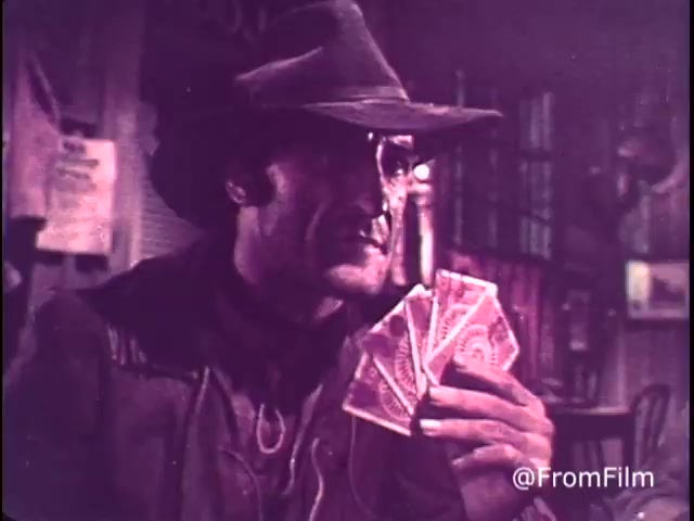 The still features a rugged character wearing a wide-brimmed hat, seated in a rustic setting. He holds a fan of playing cards in one hand, exuding a classic Western saloon vibe. The ambiance is warm, emphasizing the nostalgic feel of the 1970s. This scene is part of a playful M&M's commercial, promoting the candies with the slogan 'For good, clean fun every time.' The presence of the red M&M indicates it predates 1976, when orange was introduced.