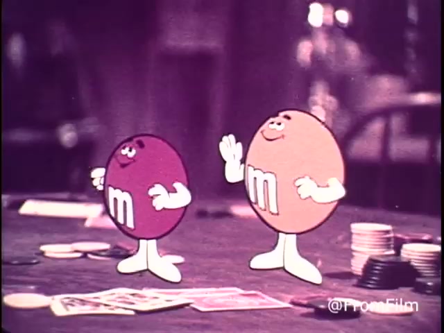 The still features two animated M&M's characters from a 1970s commercial. One is a red M&M, while the other is an orange M&M. They appear lively and playful, with cartoonish smiles and exaggerated features. The setting includes a table with playing cards and chips, suggesting a game context, aligning with the 'Go Fish' theme. The tagline 'For good, clean fun every time - M&M's chocolate candies: plain and peanut' emphasizes the playful nature of the product. The red M&M indicates that this commercial predates the 1976 changes to their color lineup.