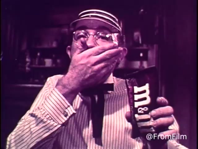 The still features an elderly man, dressed in a striped shirt and bow tie, holding a pack of M&M's in one hand while covering his mouth with the other, presumably in surprise or delight. The setting appears to be a nostalgic, cozy kitchen environment. The pack visibly contains the iconic M&M's logo, which aligns with the playful theme of the commercial. This commercial emphasizes the enjoyment of M&M's chocolate candies, reflecting the tagline 'For good, clean fun every time.' The presence of the red M&M indicates that this is from before 1976, just prior to the change in the candy's color lineup.