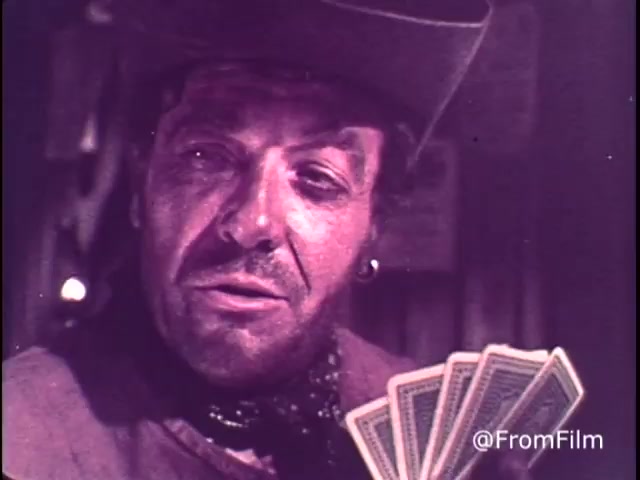 The still from the 1970s M&M's 'Go Fish' commercial features a cowboy or Western-style character, likely engaging in a card game. He has a rugged appearance with a hat and beard, and he holds a fan of playing cards. The setting appears to be an old saloon or rustic environment, aligning with common Western themes. The text emphasizes the light-heartedness of the commercial, promoting M&M's chocolate candies as a source of 'good, clean fun.' This commercial predates 1976, as indicated by the presence of the red M&M.