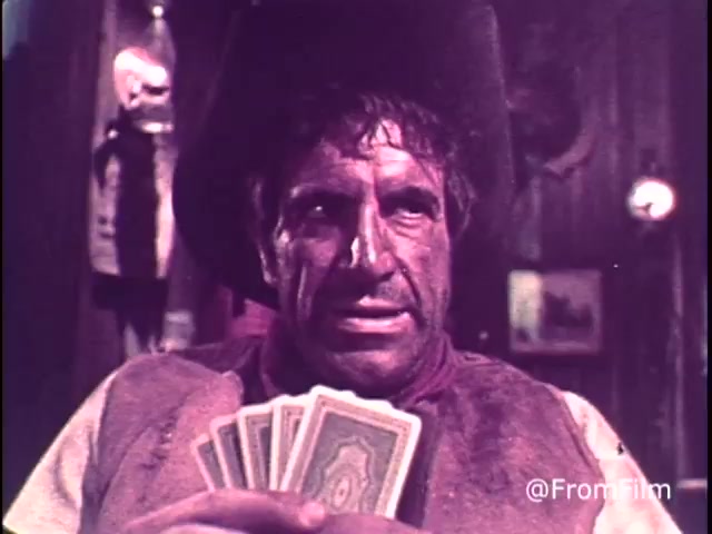 The still from the 'M&M's Go Fish' commercial features a rugged character holding a hand of playing cards. The setting appears to be a Western-themed saloon, with wooden walls and rustic decor in the background. The character, wearing a hat and looking serious, embodies the typical Western outlaw vibe. This animation likely emphasizes fun and enjoyment in a whimsical manner, fitting with the tagline about 'good, clean fun' with M&M's chocolate candies. The presence of the red M&M suggests the commercial is from before 1976.