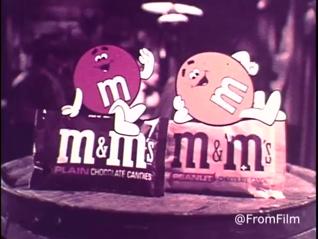 The still features two animated M&M characters, one red (representing plain chocolate) and one yellow (representing peanut). They are playfully perched on top of their respective candy packages, which display 'M&M's Plain Chocolate Candies' and 'M&M's Peanut Chocolate Candies.' The scene evokes a sense of fun and humor with bright colors and cheerful expressions, aligning with the advertising slogan, 'For good, clean fun every time.' This captures the playful essence of the 1970s M&M's commercials.