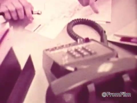 The still features a TouchTone phone prominently on a desk, showcasing its keypad. Nearby, a hand points towards a document or map, indicating a discussion likely focused on the phone's advantages. The commercial emphasizes the ease and efficiency of using TouchTone technology compared to traditional rotary phones, highlighting a modern advancement in telecommunications. The overall aesthetic seems retro, reflecting the time period of the advertisement.