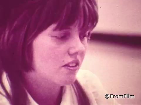 The still features a woman speaking, likely in a discussion about the benefits of TouchTone phones compared to rotary models. Her expression suggests engagement and interest in the topic. The commercial promotes the modern conveniences of TouchTone technology, highlighting its advantages in communication efficiency. The visual aesthetic reflects a vintage style, typical of mid-20th century advertising.