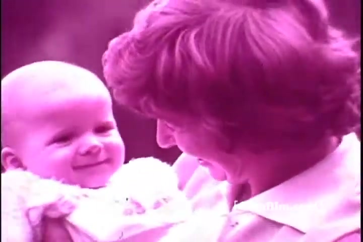 The still from the 'Exlax laxative commercial - 1970's' features a smiling woman holding a baby. Both individuals exhibit a joyful connection, with the woman looking down affectionately at the baby, who is also smiling. The image has a pinkish hue, characteristic of the era's aesthetic in advertisements. The scene likely aims to convey warmth and happiness around family, aligning with the product's messaging.