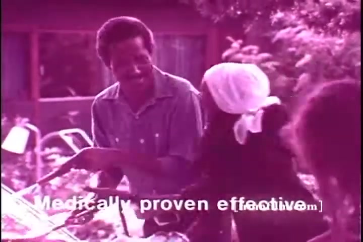 The still from the 1970s Exlax laxative commercial features a man and a woman interacting outdoors. The man appears to be smiling while engaging with the woman, who wears a headscarf. The background shows greenery and a house, creating a casual and friendly atmosphere. A text overlay states 'Medically proven effective,' emphasizing the product's credibility. The overall tone is light-hearted and approachable, typical of advertising from that era.
