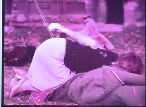 The still from the 16mm print of the 'Fuzz' trailer depicts two figures on the ground in a dynamic posture, suggesting action or tension. The scene features muted colors typical of older film formats, with scratches and imperfections evident in the print, conveying a vintage aesthetic. The background appears blurred, drawing focus to the foreground figures. The overall atmosphere hints at a comedic or suspenseful moment, characteristic of the film's genre.