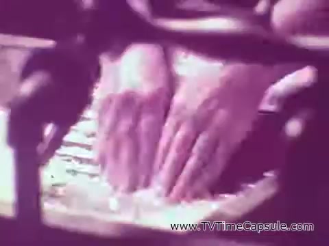 The still features two hands pressing down on a shiny surface, possibly a kitchen or cleaning product. The focus is on the hands, with a blurred background that suggests a commercial setting. The lighting gives the image a slight purplish hue, emphasizing the action taking place.