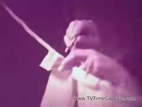 The still from the 'ScotTowels Commercial' features a close-up of a hand holding a roll of paper towels. The color has a slight purple hue, giving it a vintage feel. The focus is on the hand, emphasizing the action of pulling a towel from the roll, suggesting practicality and ease of use. The overall aesthetic reflects a classic advertising style.