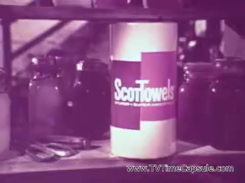 The still features a cylindrical container of ScottTowels prominently displayed on a shelf. The packaging is simple, with the brand name clearly visible. In the background, there are jars and utensils, suggesting a domestic or kitchen setting. The overall tone is reminiscent of mid-20th century advertising, focusing on practical household products.