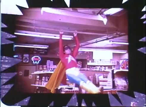 The still features a vibrant scene from the film 'Hero at Large,' showcasing a comedic moment with the character dressed as a superhero. The character, wearing a bright red costume with a yellow cape, appears to be swinging or hanging from a ceiling in a convenience store setting. The background includes shelves filled with products and a neon sign, adding to the playful, lighthearted tone typical of 1980s comedies. The edges of the image are framed with a stylized design, emphasizing its promotional nature.