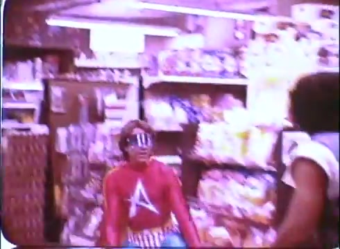 The still from the 16mm trailer of 'Hero at Large' features a vibrant scene set in a grocery store. In the foreground, a character in a colorful superhero costume, complete with a red top and blue pants, is striking a playful pose. He wears oversized sunglasses, enhancing the comedic tone of the moment. Shelves filled with various snacks and products are visible in the background, contributing to the lighthearted, everyday setting of the film. This promotional image captures the whimsical spirit of the comedy starring John Ritter.