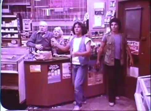 The still from 'Hero at Large' features a scene set inside a store, where four characters—two men, a woman, and an older man—are reacting with surprise or concern. One of the men is holding a weapon, indicating a tense situation. The characters are dressed in casual attire typical of the late 1970s and early 1980s. The interior of the store has various items and signs visible, including a prominently placed 'OPEN' sign. The overall tone suggests a mix of comedy and drama, characteristic of the film's premise.