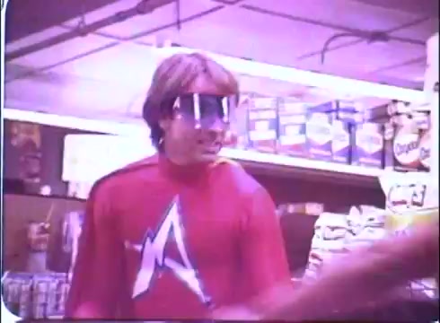 The still from 'Hero at Large' features a young man, played by John Ritter, wearing a bright red superhero costume with a prominent white 'A' emblem. He also has distinctive oversized glasses that add a whimsical touch to his character. The scene appears to be set in a supermarket, with shelves stocked with various products in the background, creating a humorous contrast between the superhero persona and an everyday environment. The overall tone conveys a lighthearted and comedic vibe typical of the film.