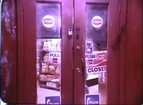 The still features a pair of large, glass double doors leading into a store, showcasing a view of shelves filled with various products inside. The doors are adorned with signs indicating 'PULL' and 'PUSH,' alongside a prominent 'CLOSED' sign. The ambiance appears nostalgic, typical of the early 1980s, reflecting the comedic and lighthearted tone of 'Hero at Large,' which stars John Ritter as an ordinary man who finds himself in heroic circumstances. The overall visual captures a moment that sets the stage for the film's humorous plot.