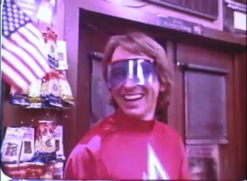 The still features John Ritter in a vibrant red superhero costume, complete with a bold letter 'A' on the chest. He wears oversized blue sunglasses and has a cheerful smile, suggesting a light-hearted and comedic tone. The background includes snacks and an American flag, hinting at a playful and patriotic setting. This image captures the whimsical spirit of 'Hero at Large,' emphasizing Ritter's comedic charm.
