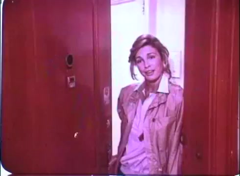 The 16mm still is a promotional image from the 1980 film 'Hero at Large.' It features a woman standing at an apartment door, smiling softly towards the camera. She is dressed in a light-colored jacket, and the warm, slightly faded colors of the film print evoke a nostalgic feel typical of the era. The setting appears to be a cozy, domestic space, aligning with the film's comedic and light-hearted themes.