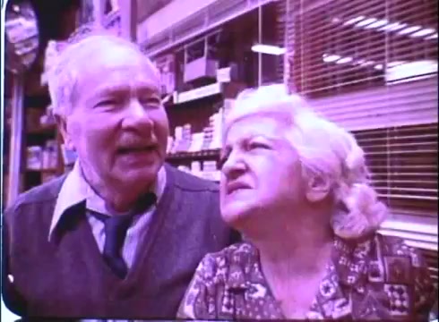 The still from the 'Hero at Large' movie promos features an older couple, with the man laughing and the woman looking at him with a playful expression. They appear to be in a store, likely engaged in a light-hearted conversation. The scene captures a charming, comedic moment, typical of the film's lighthearted tone. The image is presented in a vintage style, characteristic of a 16mm print, with a slightly faded color palette.