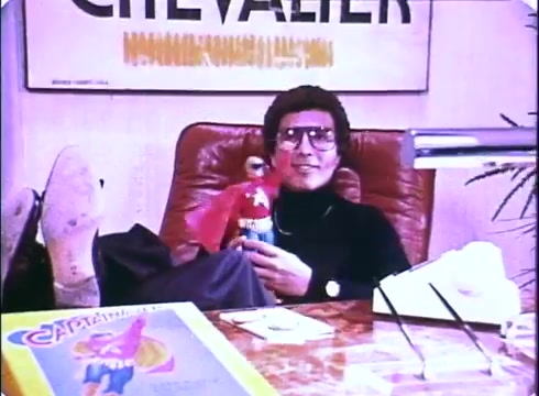 The still from the 'Hero at Large' movie promos features a relaxed John Ritter sitting at a desk, with his feet up. He holds a toy figure resembling a superhero, showcasing a playful and lighthearted vibe. The background includes promotional materials, likely related to the film, emphasizing its comedic essence. The colors and style reflect the period's aesthetic, characteristic of early 1980s cinema.