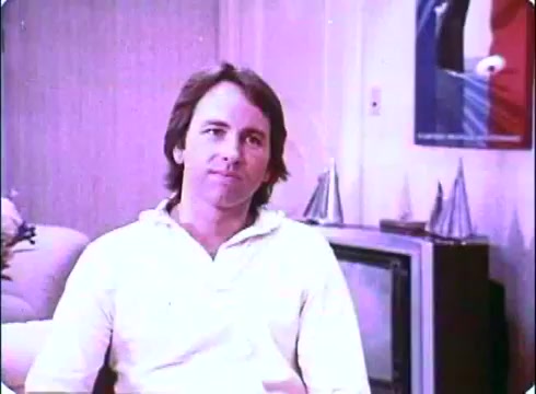 The still from the 'Hero at Large' movie promos features John Ritter seated in a casual white sweatshirt, looking contemplative. The environment appears cozy, with a sofa and a vintage television in the background. The walls have minimal decor, and a poster is visible, hinting at the film's promotion. The overall mood seems relaxed, reflecting the comedic tone of the film.