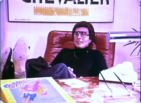 The still from the 'Hero at Large' movie promos features a character seated casually in an office setting, reclining in a leather chair with their feet up on a desk. The individual is wearing glasses and a black outfit, exuding a relaxed demeanor. On the desk, there is a magazine or comic titled 'Captain...', hinting at a superhero theme. The background includes a poster with the word 'CHEVALIER,' contributing to the overall comedic and laid-back atmosphere of the film's promotion.